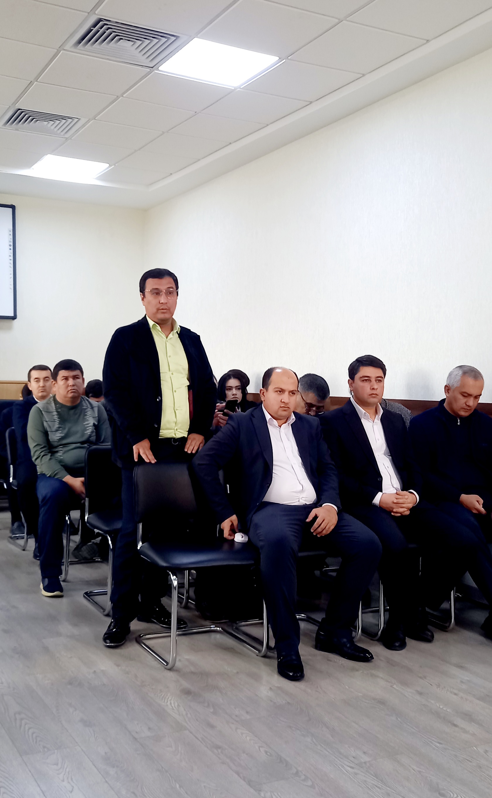 A group of men in suits and ties sit in black chairs in a room with light-colored walls and ceiling, facing forward.