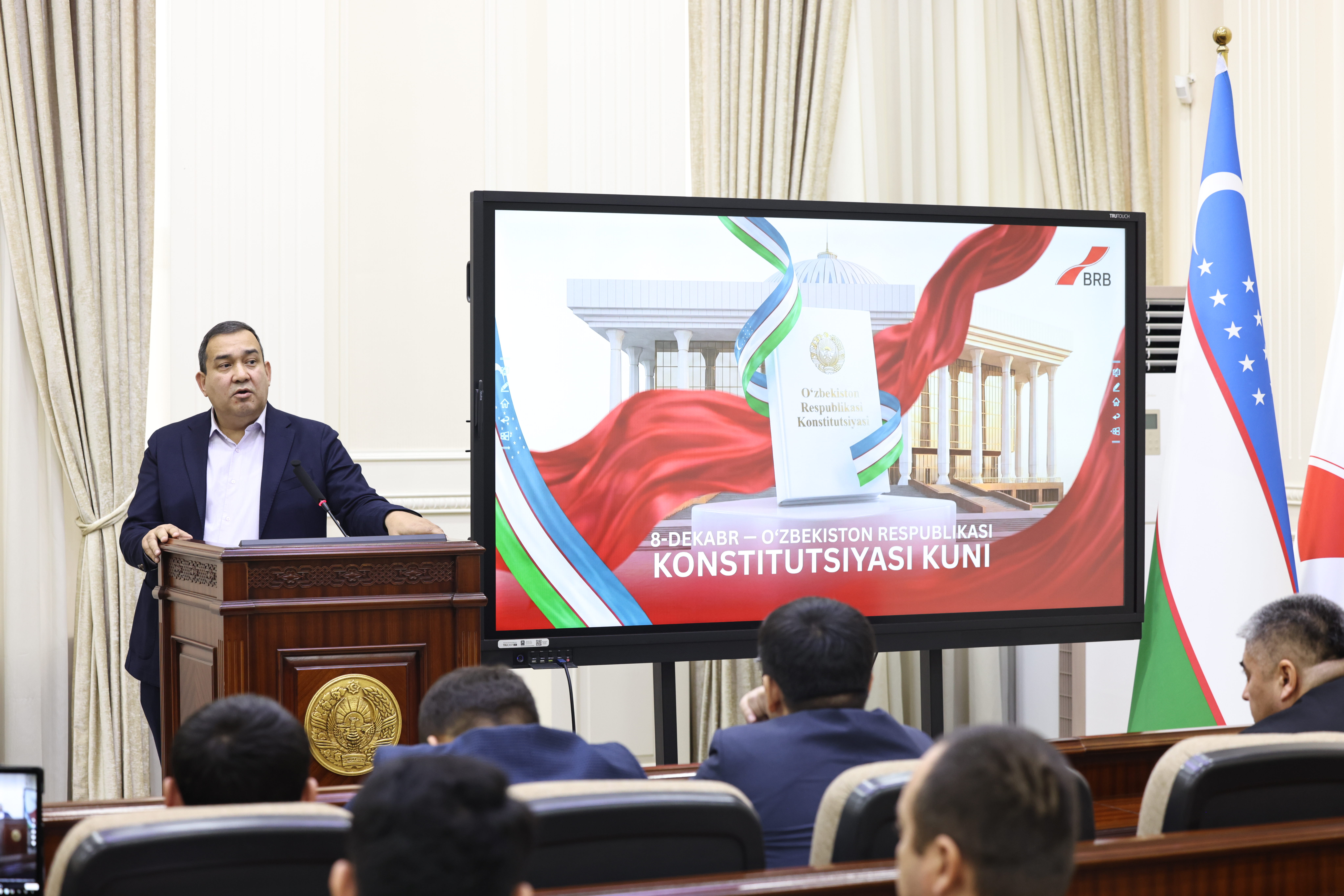 A man in a suit speaks at a podium in front of a large screen displaying a government building and text, with a group of people seated in front of him.