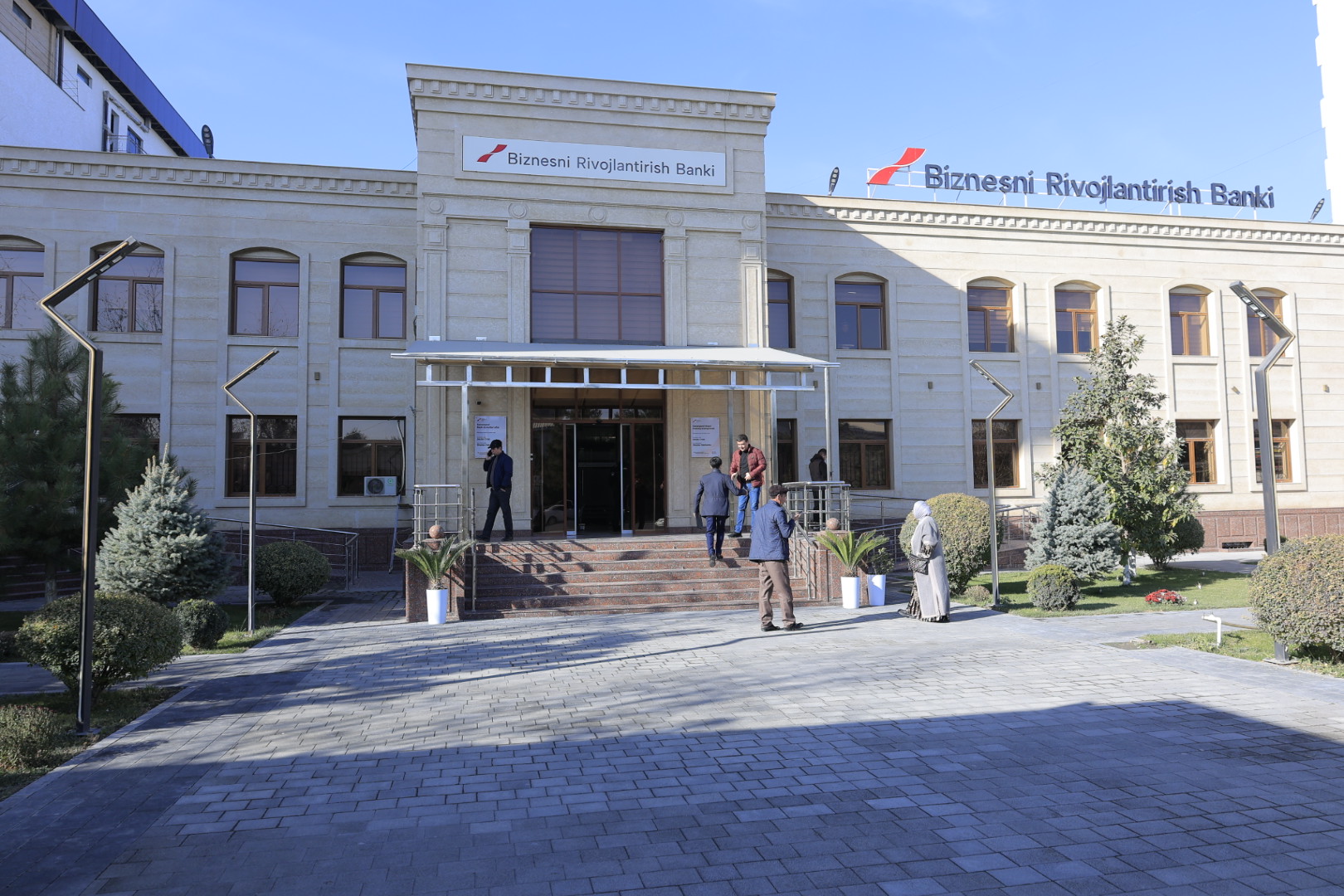A beige building with arched windows and a sign reading "Biznesni Rivojlantirish Bank" stands under a clear blue sky, with three people near the entrance.