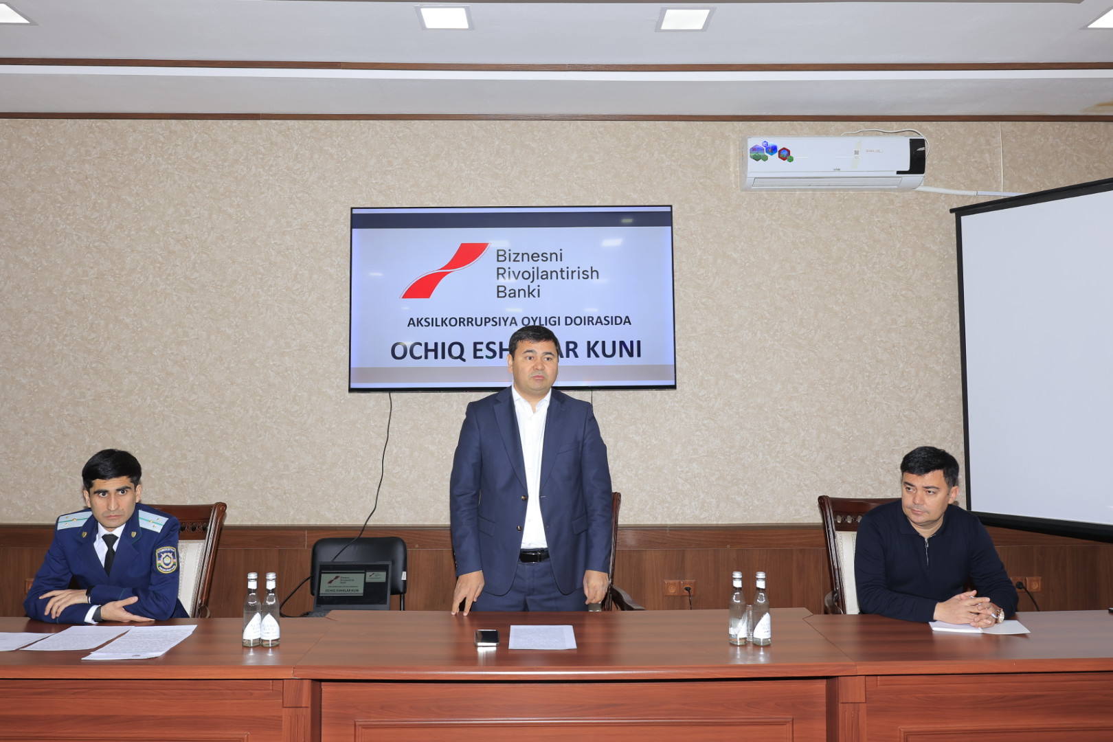 Three men in suits sit at a wooden table in front of a screen displaying "Biznesni Rivojlantirish Bank" and "OCHIQ ESKR KUNI", with a projector screen to the right.