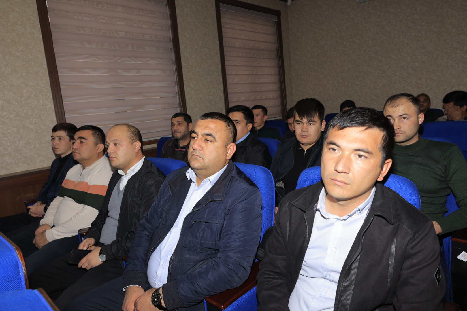 A group of men in dark jackets and light shirts sit in blue chairs, facing forward in a room with light-colored walls and blinds on the windows.