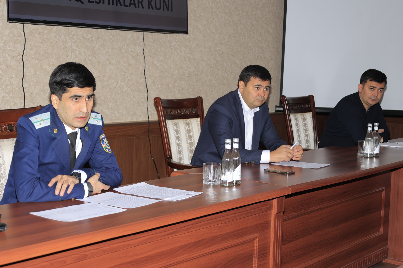 Three men in suits and ties sit at a wooden table with papers and water bottles, facing a screen displaying text.