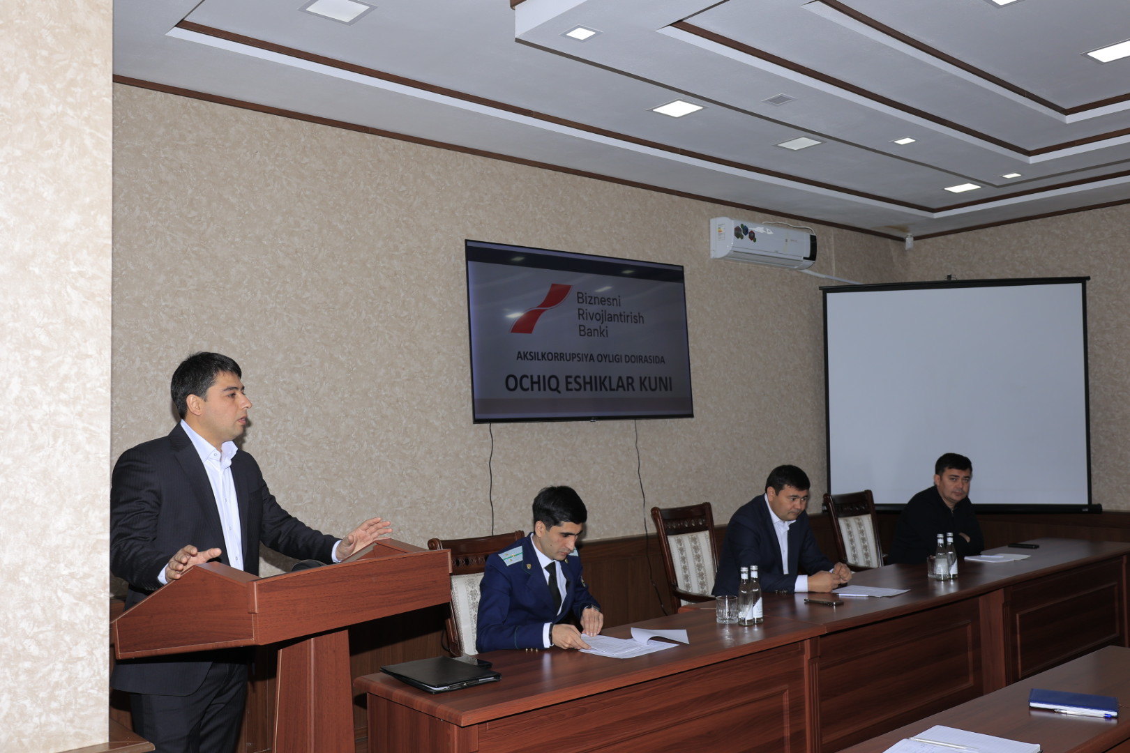 A man in a suit speaks at a podium while three others sit at a table in a conference room with a projector screen and wall-mounted sign.