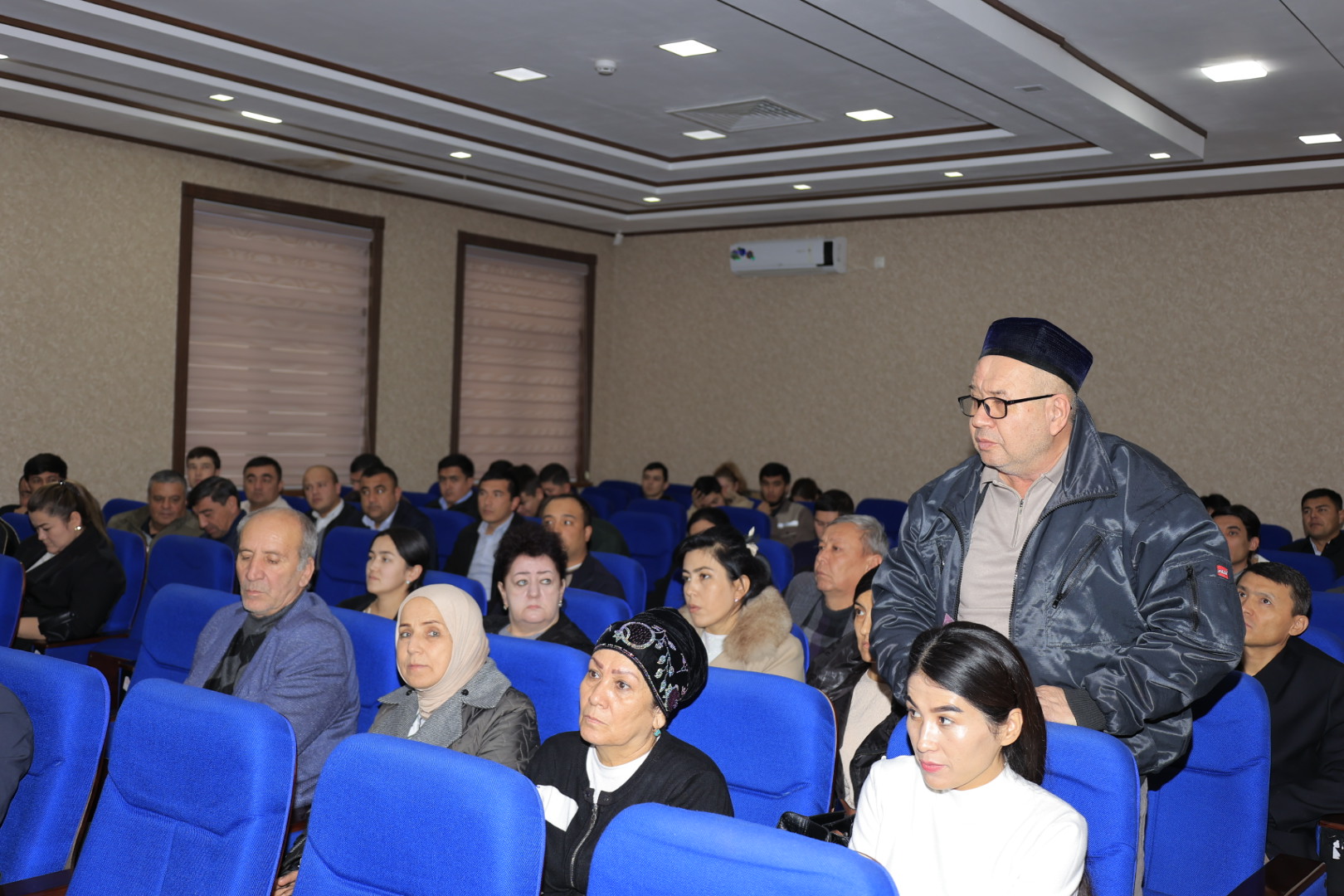 A man in a dark jacket and cap stands at the front of a blue-cushioned auditorium, facing an audience seated in rows of blue chairs.