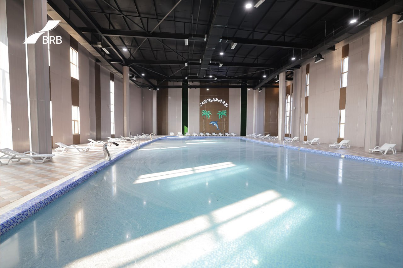 An indoor swimming pool with light blue water, white lounge chairs, and a high ceiling with lights and structural beams, with a mural of palm trees and a dolphin on the wall.