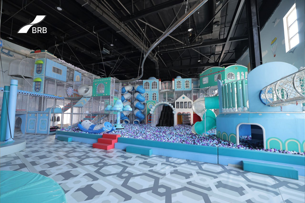 An indoor playground with blue and white structures, slides, climbing elements, and a blue-floored area with blue pebbles, under a black ceiling with exposed pipes and ductwork.