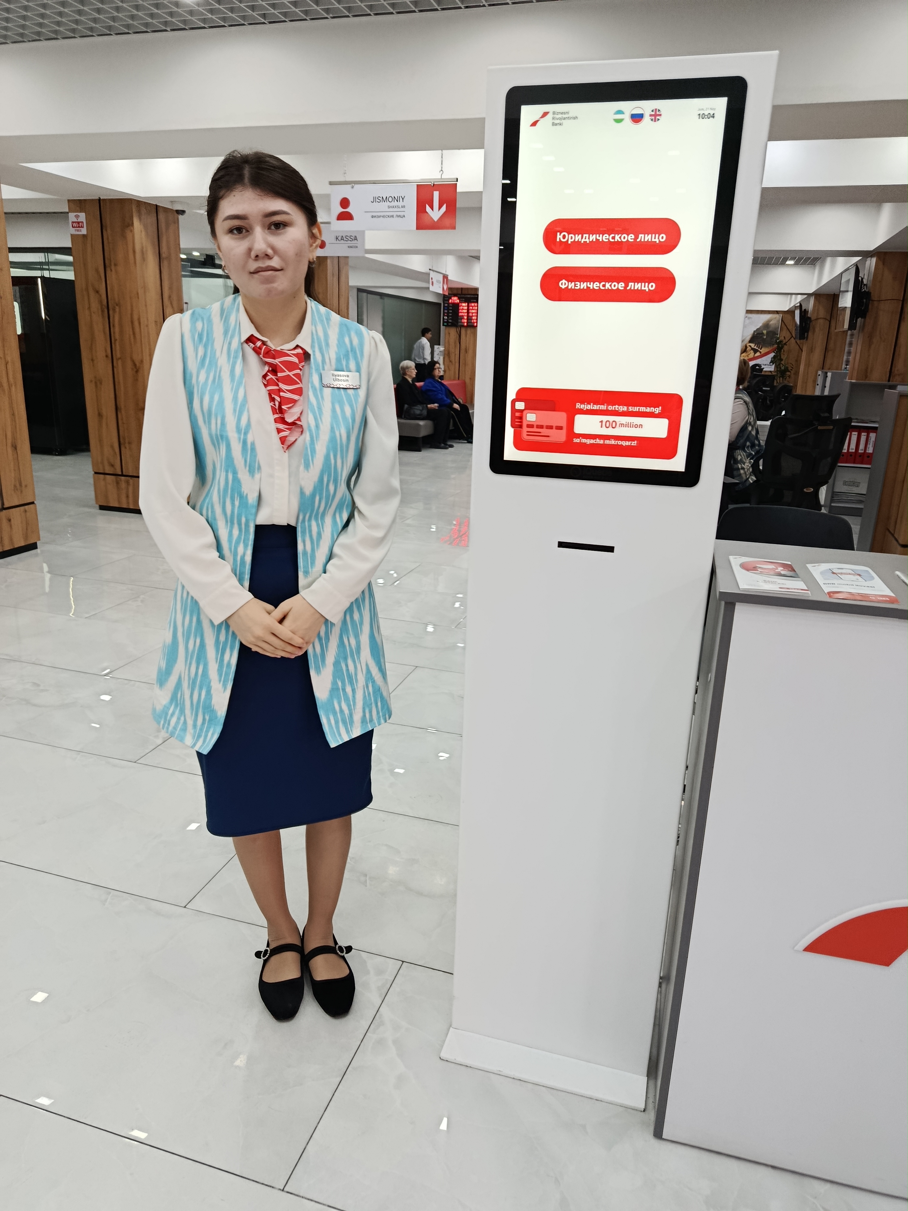 A woman in a light blue and white blazer, dark blue skirt, and black shoes stands next to a white kiosk displaying red text and buttons in a hallway.