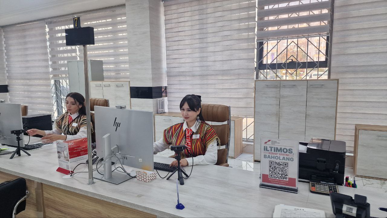 Two women in patterned shirts and ties sit at a light-colored desk with computer monitors, phones, and office supplies, in front of blinds covering a window.