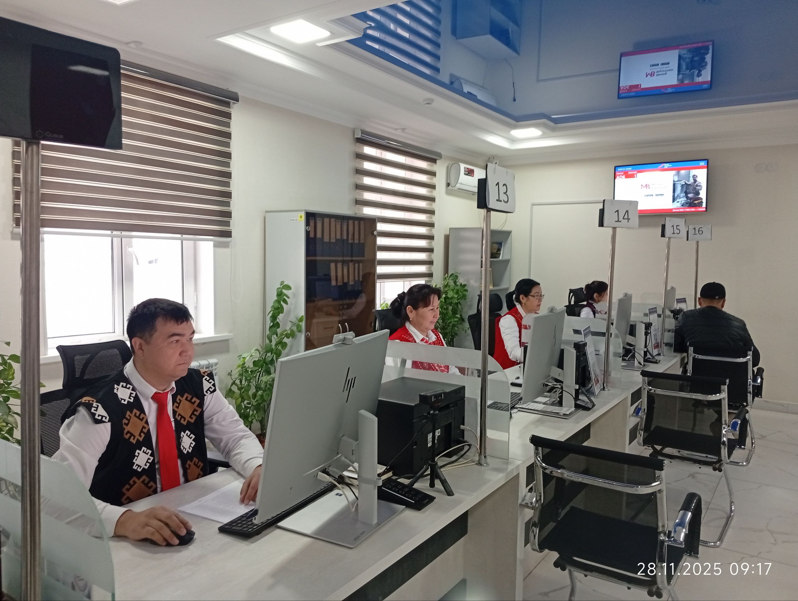 Four people sit at desks with computer monitors and chairs in a room with blinds on the windows and TVs mounted on the walls.
