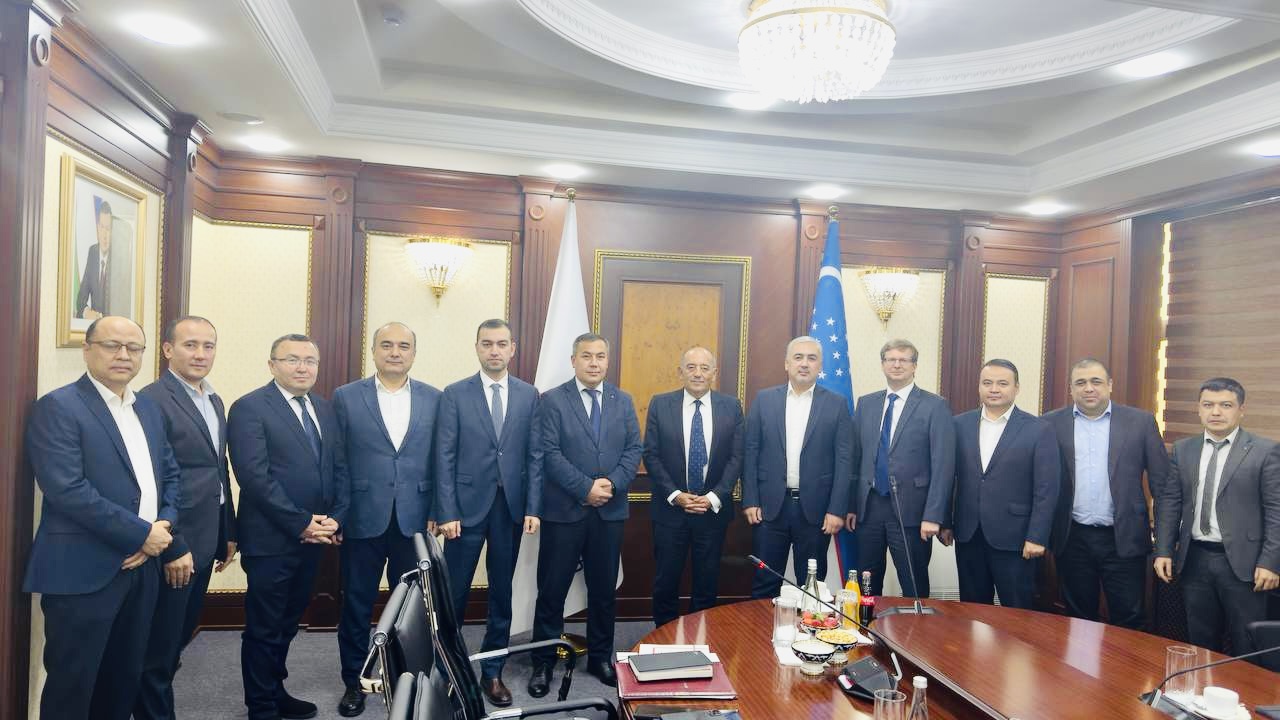 A group of men in suits stand in a formal setting with wood-paneled walls, a chandelier, and a flag.