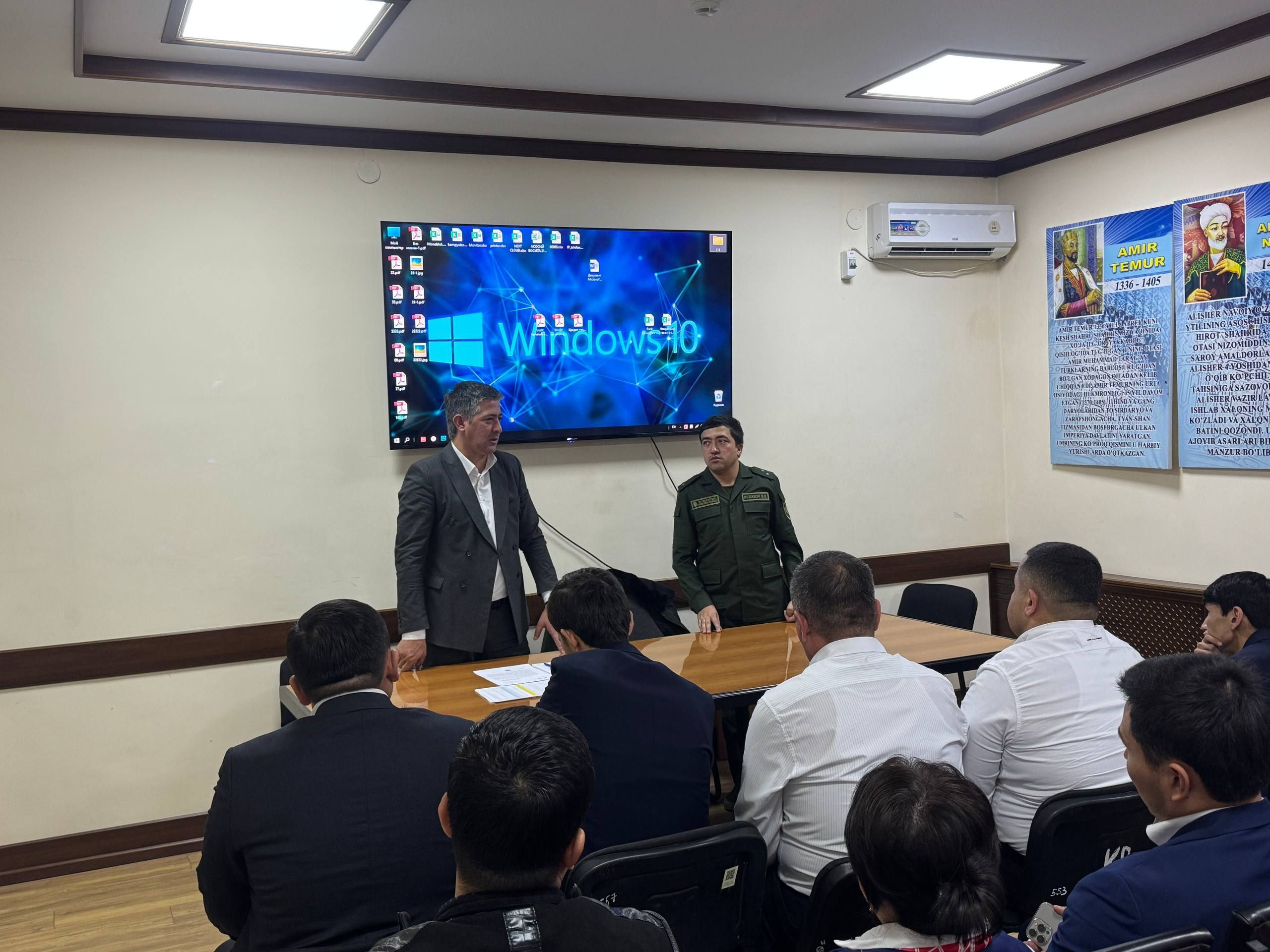 A man in a suit stands before a large screen displaying "Windows 10," addressing a group of people seated around a table in a room with beige walls and wooden trim.