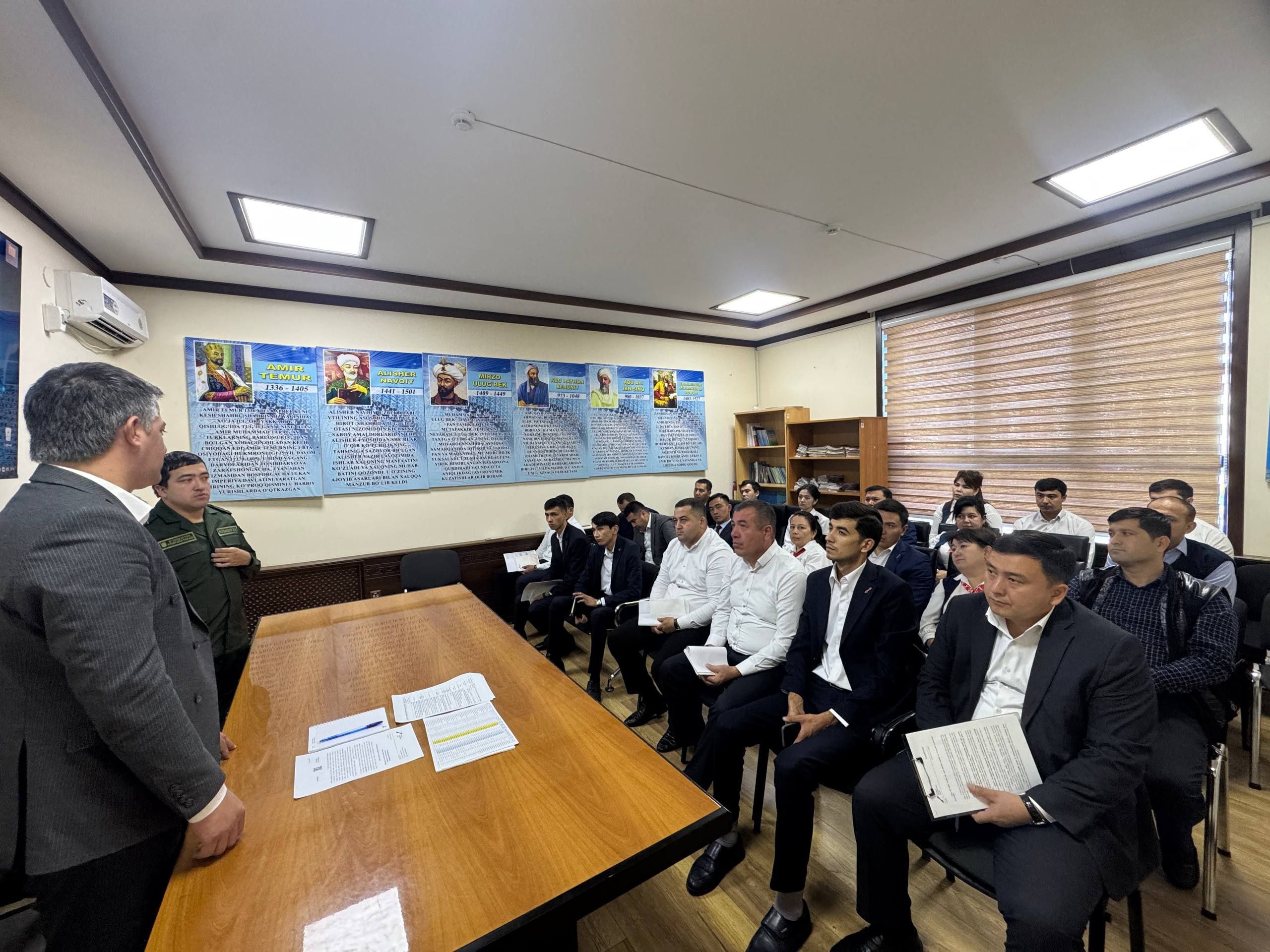 A group of people in white shirts and dark suits sit in chairs facing a man in a dark suit at a long wooden table with papers and documents.