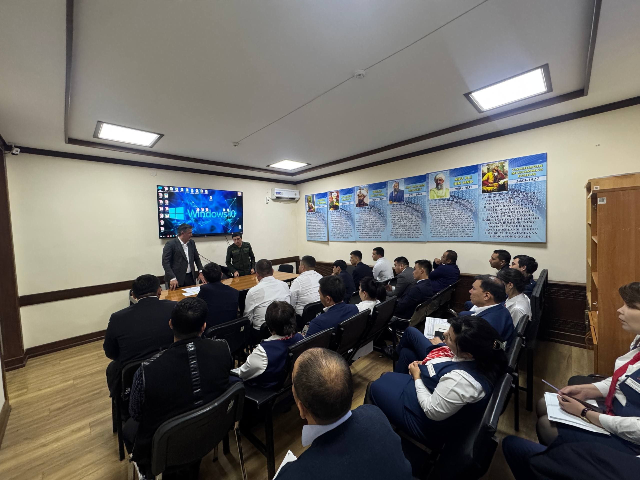 A teacher stands at the front of a classroom, addressing students seated in rows, with a large screen displaying a blue image and text on the left wall and a blue banner with images and text on the right wall.