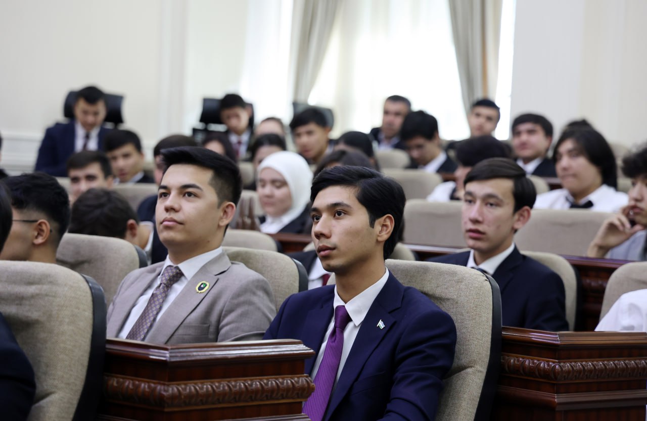 A group of young men in suits and ties sit in rows on wooden benches, facing forward in a formal setting.