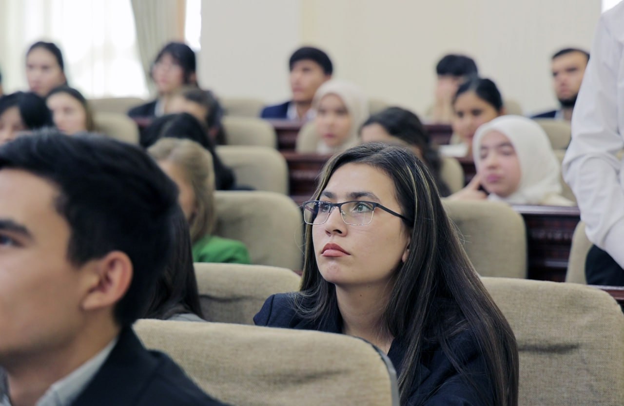 A classroom of students in beige chairs faces forward, with two students in the foreground wearing glasses and dark jackets.
