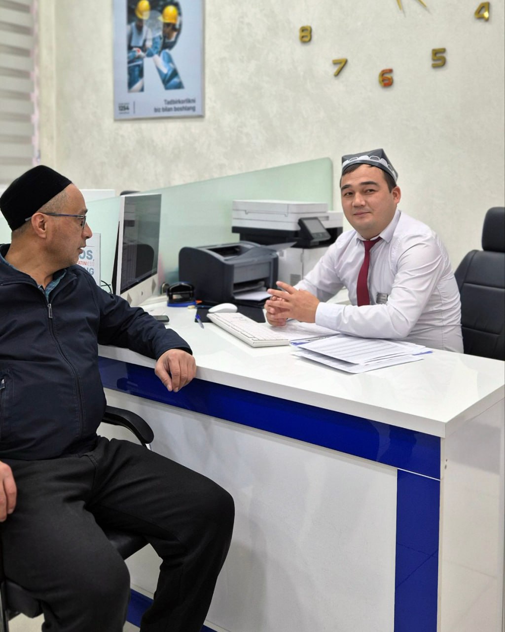 Two men sit at a white desk with blue trim; one in a black jacket and glasses, the other in a white shirt and red tie, with a computer and printer behind them. A poster and clock are visible on the wall.