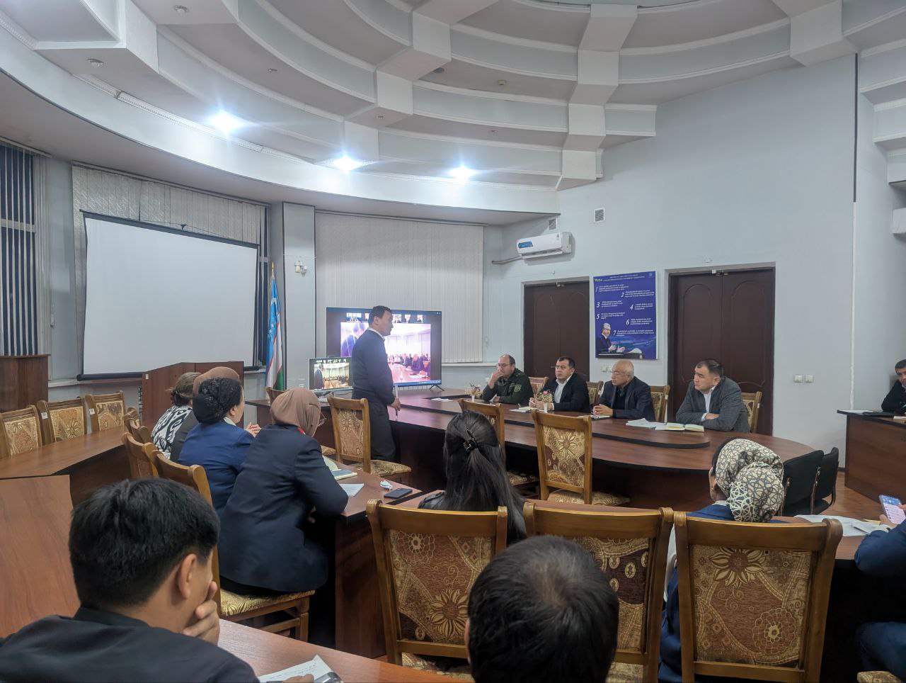 A conference room with a curved white ceiling, projector screen, and brown chairs with patterned upholstery hosts a meeting with a speaker at the front.