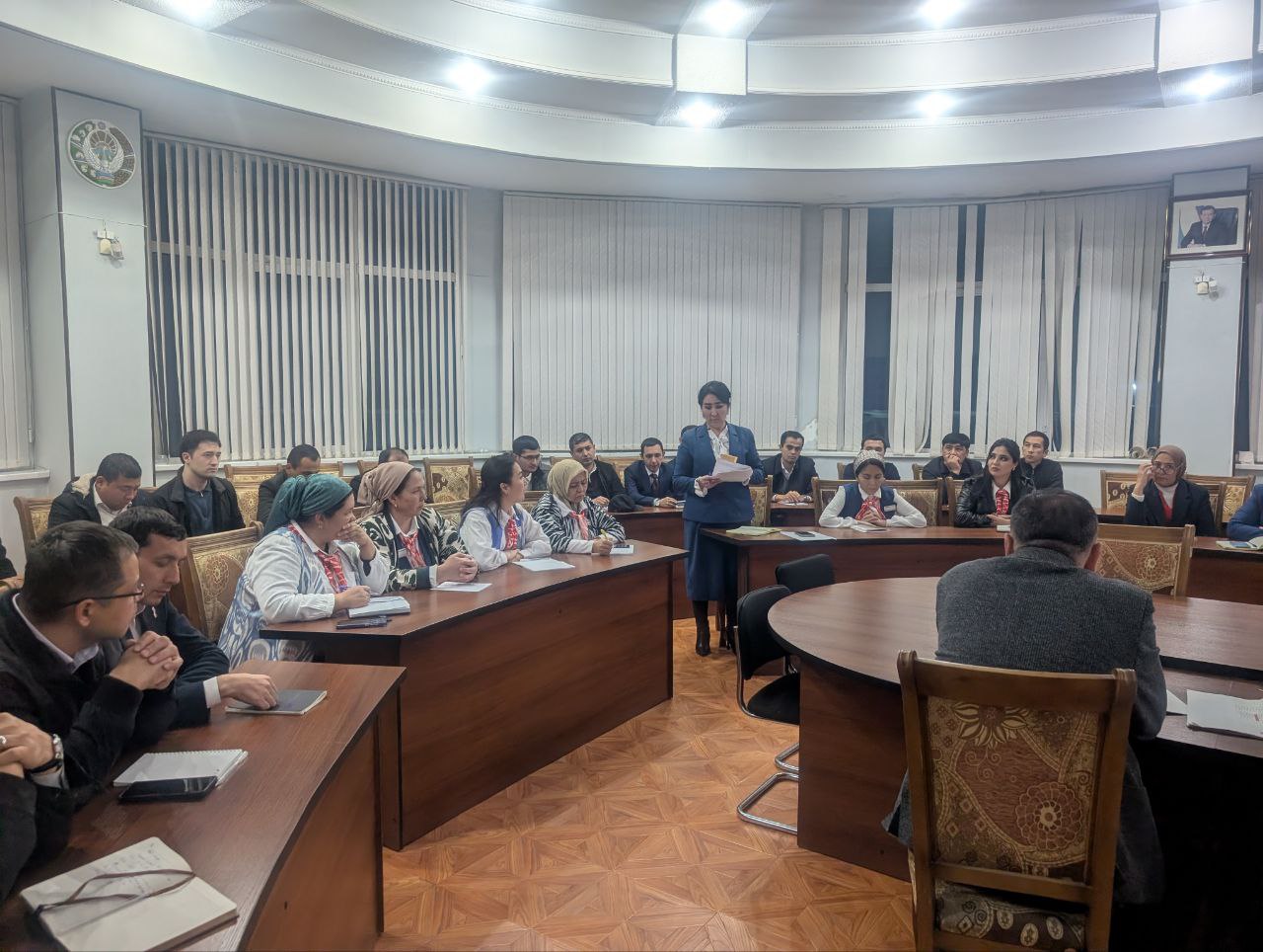 A formal meeting or conference room with wooden desks, chairs, and large windows with blinds. A man in a dark suit stands at the front addressing the group.