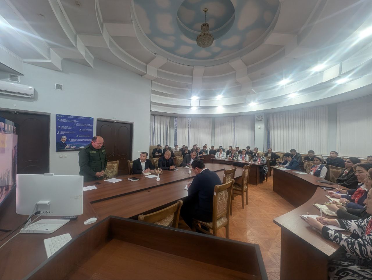 A group of people sit around a long wooden table in a room with a curved ceiling and ceiling fan, facing a man speaking at a podium.
