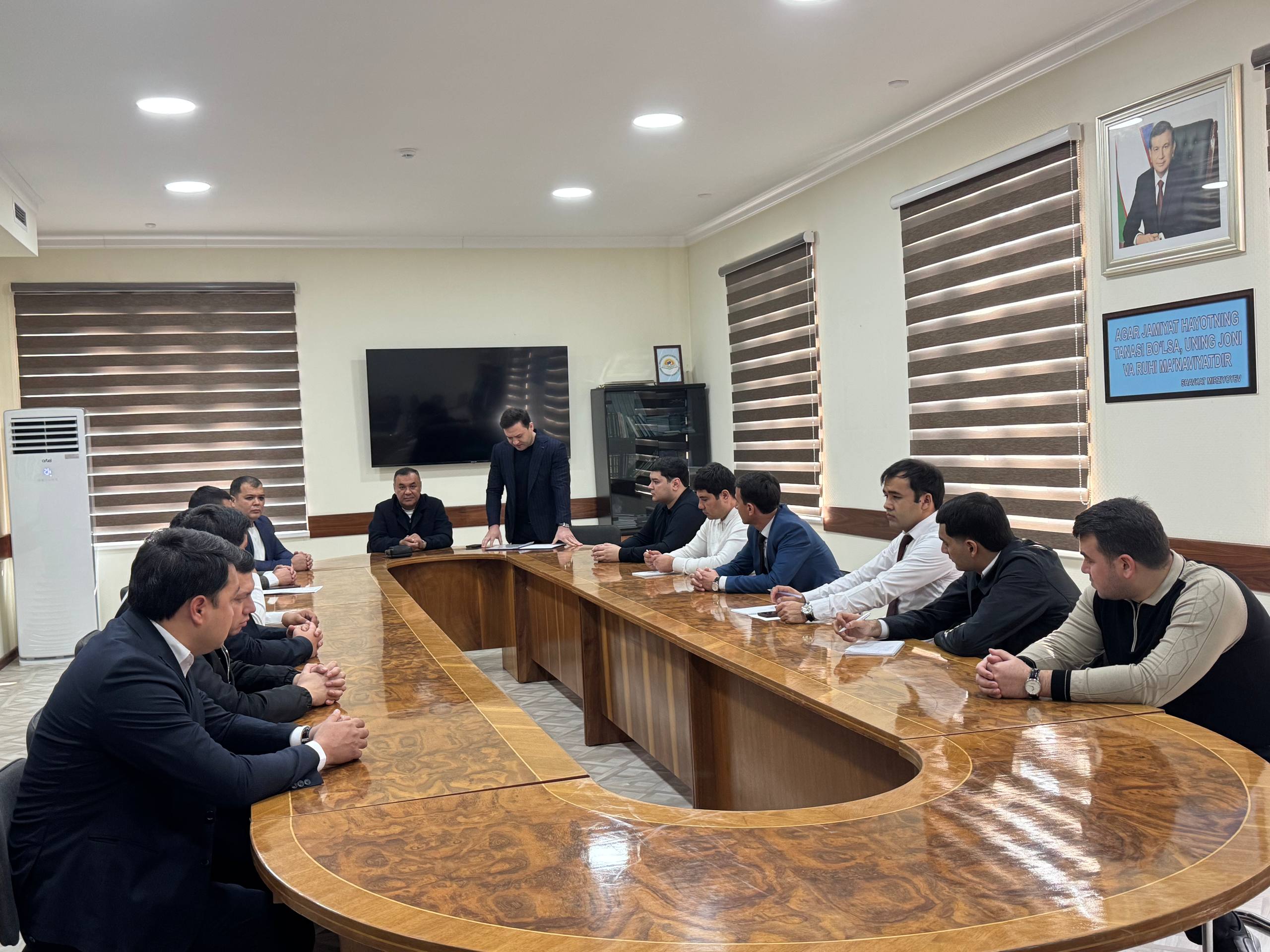 A group of people in business attire are seated around a large, curved wooden table in a meeting room.