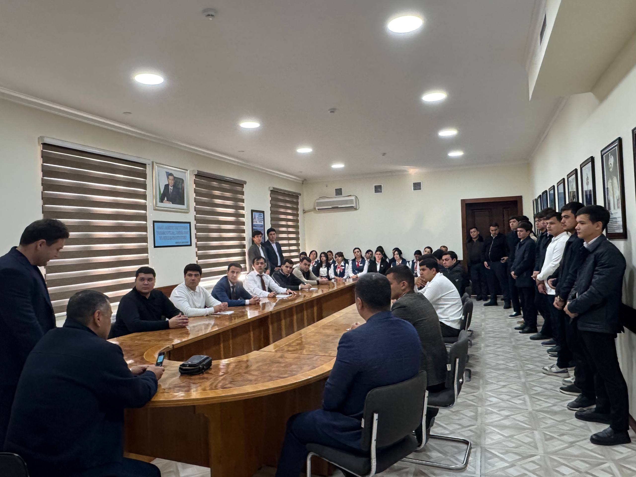A group of people in business attire are seated around a long wooden table in a meeting room with blinds on the windows and framed pictures on the wall.