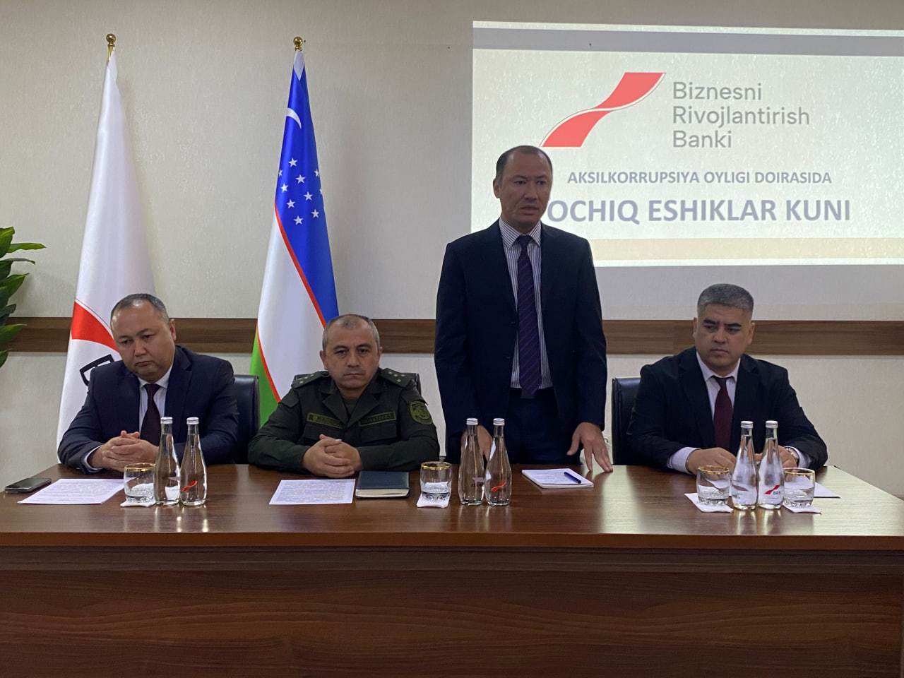Four men in suits and ties sit at a wooden table with papers and water bottles, in front of a projector screen displaying the Biznesni Rivojantirish Bank logo.