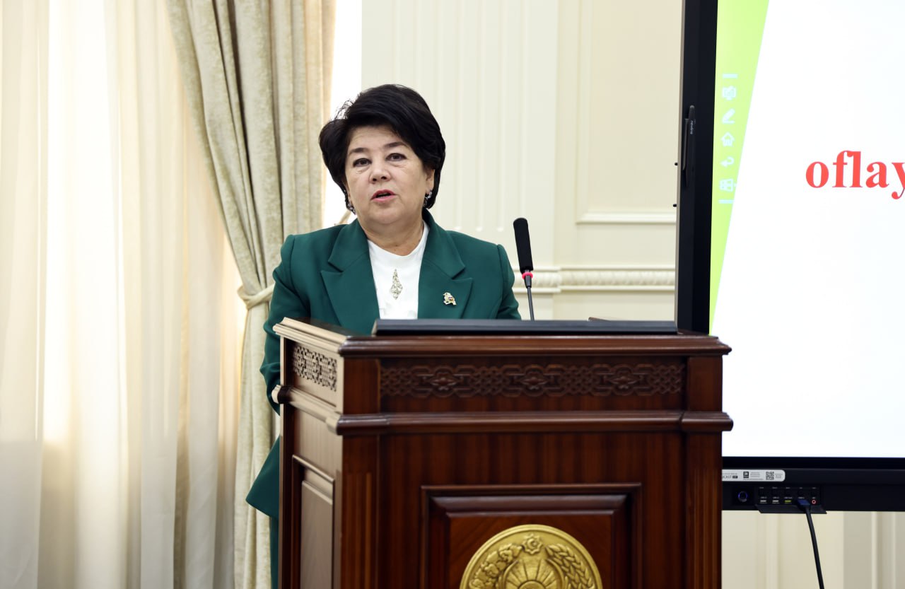 A woman in a green blazer stands behind a wooden podium with a gold emblem, speaking into a microphone. A projector screen displaying "oflay" is visible to her right.