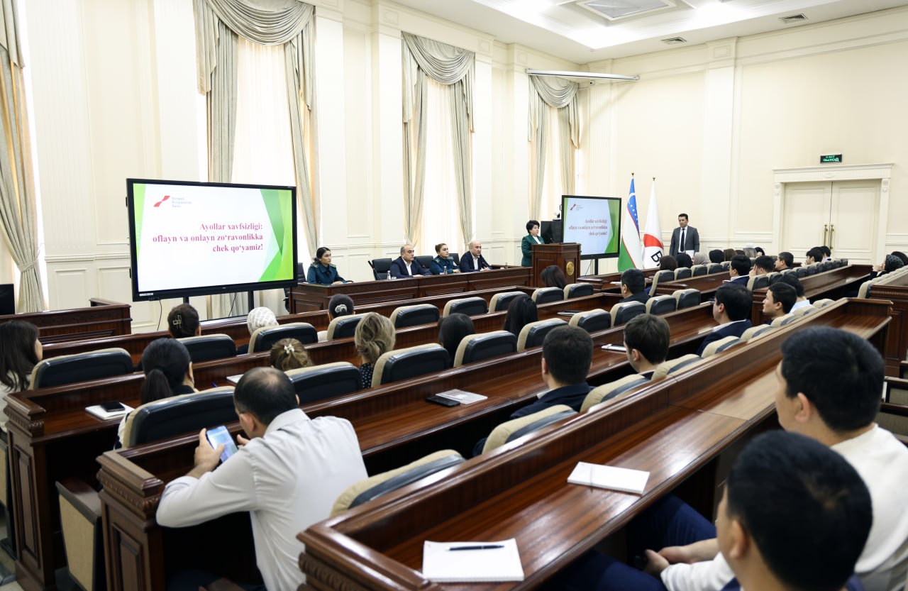 A conference room with rows of wooden desks and large screens displays a presentation in French, with attendees seated and observing.