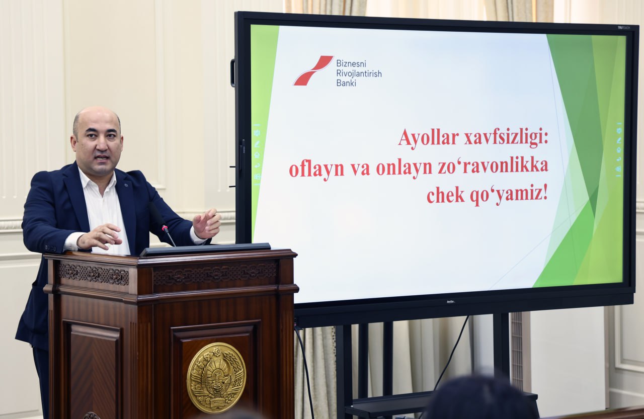A man in a suit stands at a podium, speaking to a large screen displaying text in red and white against a light green and white background.