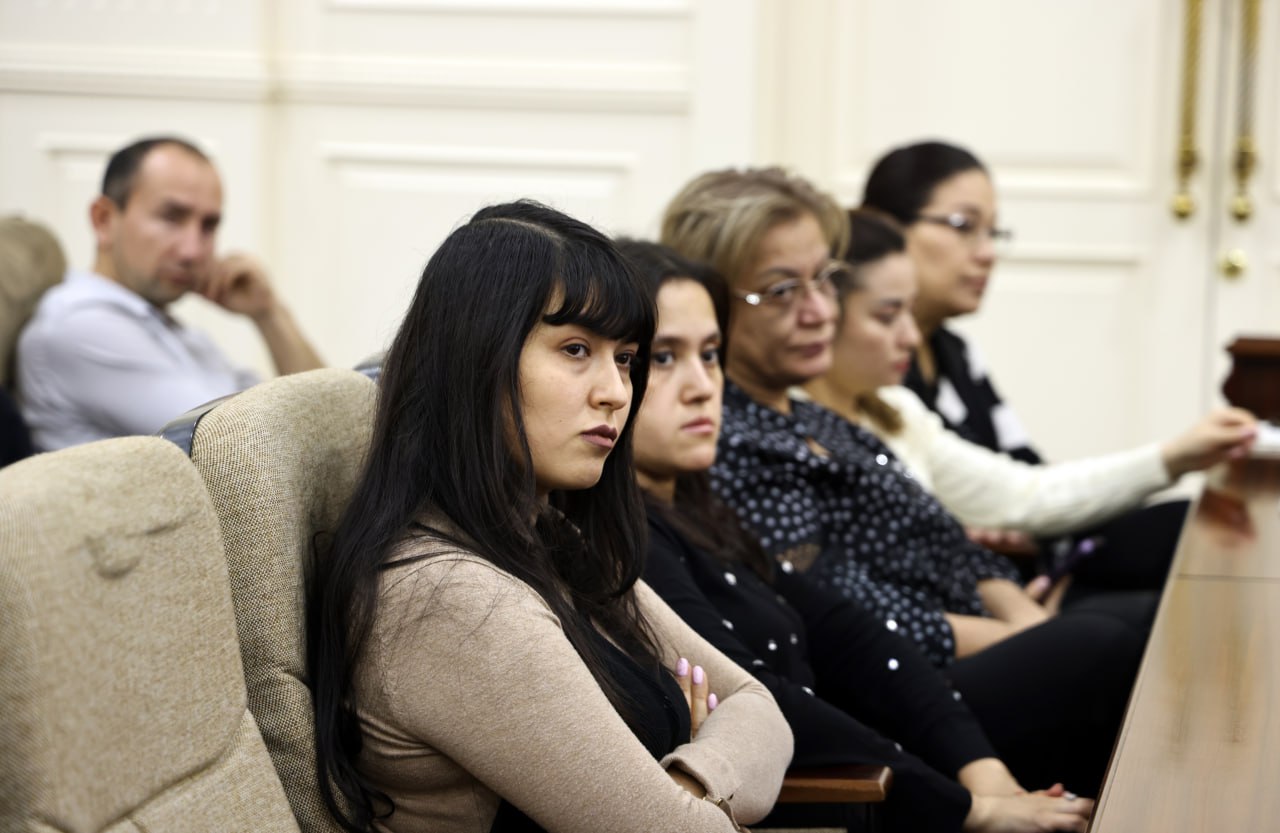 Five women with dark hair and glasses sit in a row, facing the same direction, with two men visible in the background.