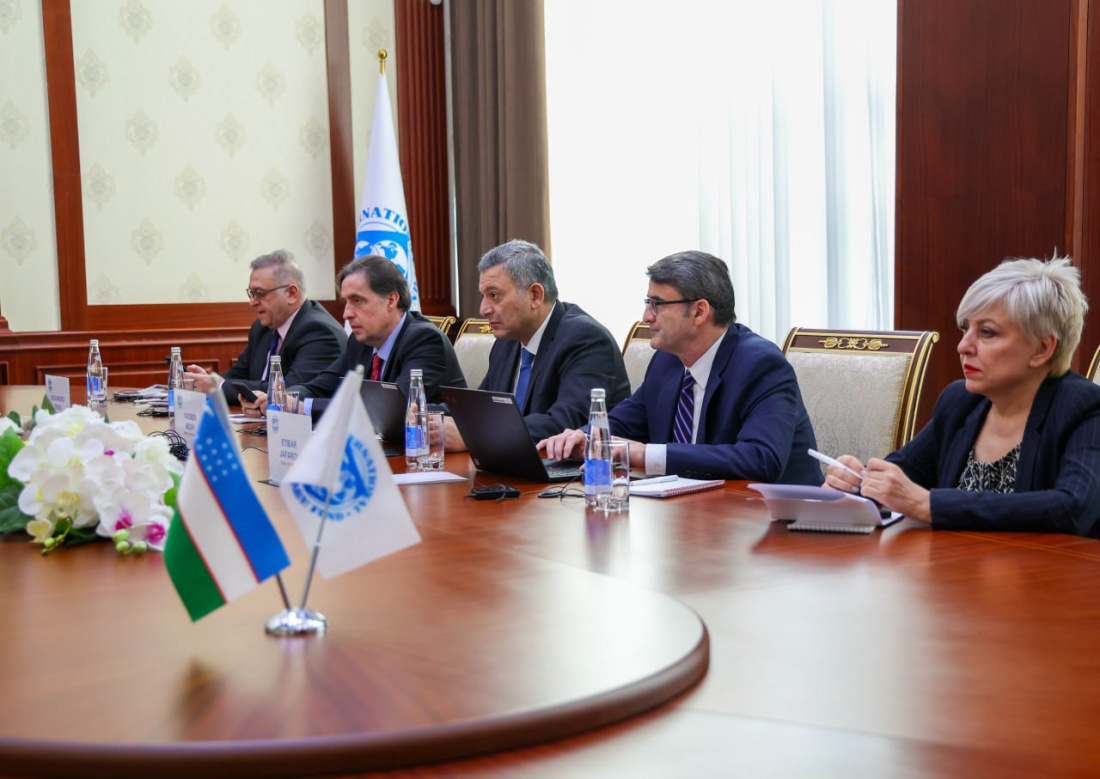 Five men in suits sit around a wooden table with laptops and water bottles, flanked by small national flags.